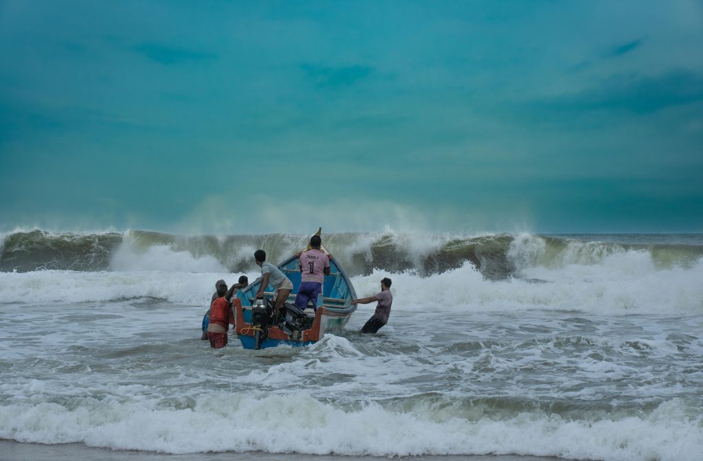 A group of fishermen launch their boat amidst powerful ocean waves under a blue sky.