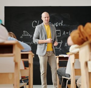 Bald teacher explaining geometry on blackboard to attentive students in classroom.