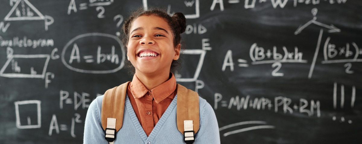 Smiling girl with backpack in a classroom, standing in front of a chalkboard with math formulas.